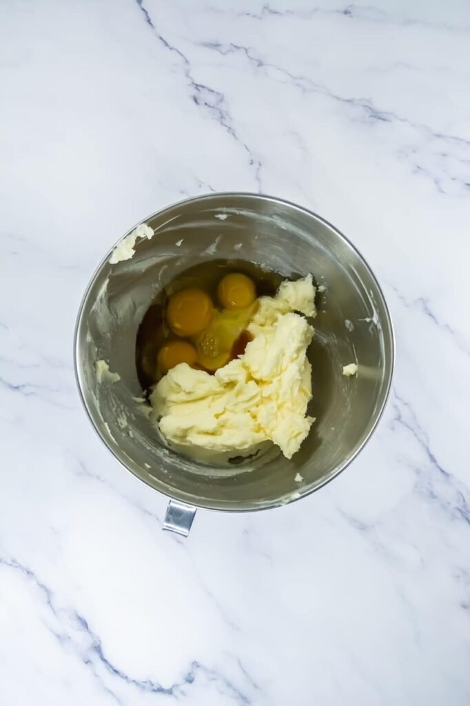 Overhead view of a stand mixer bowl with softened butter, cracked eggs, and vanilla before mixing, showing the early stage of making Crumbl churro cookie dough on a white marble surface.