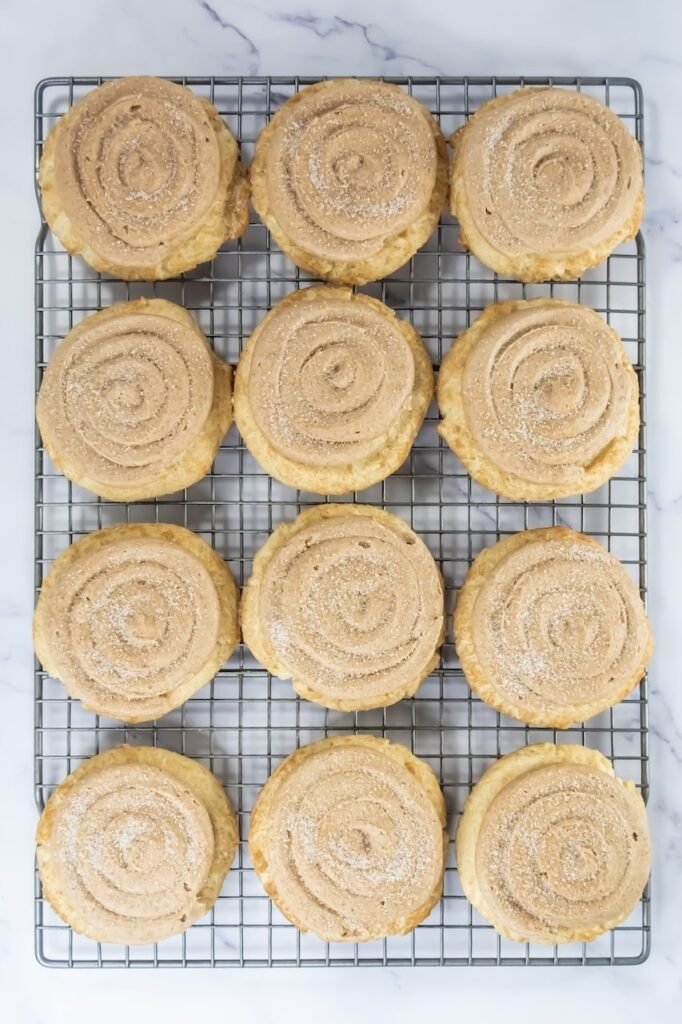 Overhead view of Crumbl churro cookies cooling on a wire rack, each topped with a spiral of cinnamon buttercream frosting and lightly sprinkled with cinnamon sugar.