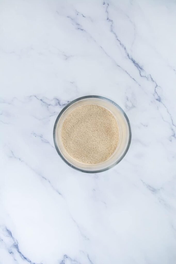 Overhead view of a small glass bowl filled with cinnamon sugar mixture on a white marble surface, prepared for coating dough before baking.