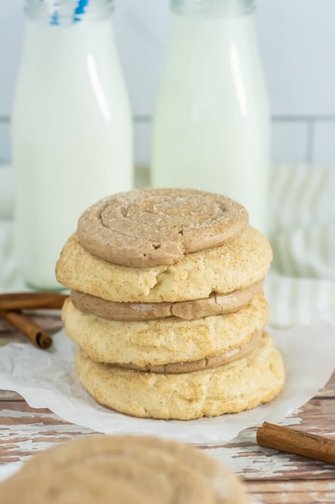 Stack of thick Crumbl churro cookies with cinnamon buttercream frosting between layers, styled on parchment paper with cinnamon sticks and bottles of milk in the background.