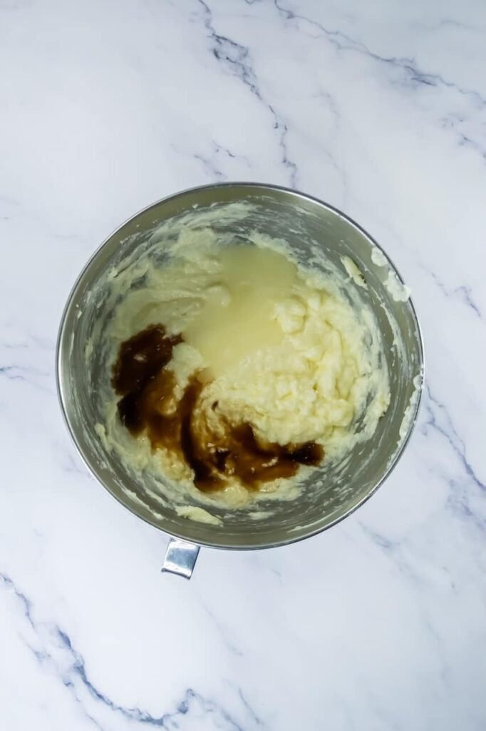 Overhead view of a stand mixer bowl with creamed butter and sugar, showing egg whites, vanilla extract, and cake flavoring added on top before mixing. This image shows the wet ingredients being incorporated to create a smooth, light base for confetti cake cookie dough.