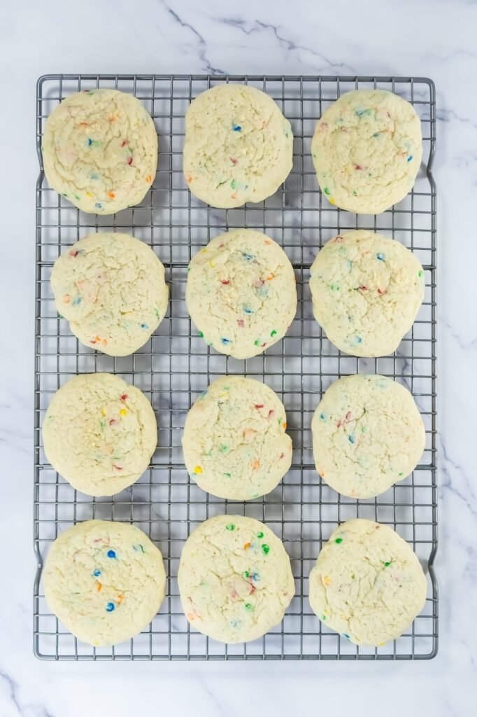 Overhead view of freshly baked desserts cooling on a wire rack, showing thick, pale cookies with soft centers and colorful sprinkles throughout. This image shows the cookies after baking and before frosting, demonstrating proper doneness and texture.