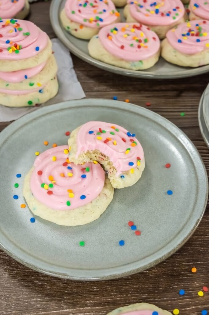 Close-up view of two confetti cake cookies on a light-colored ceramic plate, topped with smooth pink cream cheese frosting and colorful round sprinkles. One cookie has a large bite taken out, clearly showing the soft, fluffy, cake-like interior with small sprinkles baked throughout the crumb. Loose sprinkles are scattered on the plate and wooden table, emphasizing the texture and softness of the cookies and the thickness of the frosting swirl.