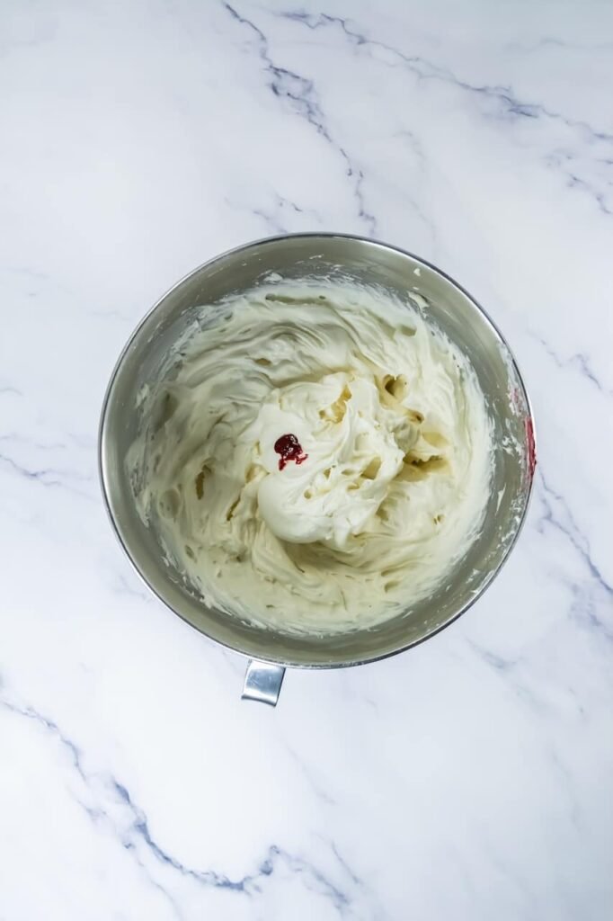 Overhead view of whipped cream cheese frosting in a stand mixer bowl, showing a smooth, fluffy texture with a small amount of pink food coloring added on top. This image shows the frosting at the final mixing stage before piping.