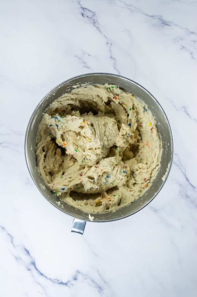 Overhead view of finished dough in a stand mixer bowl, showing a thick, soft texture with colorful sequin sprinkles evenly mixed throughout. This image represents the completed cookie dough after folding in sprinkles and before scooping and baking.