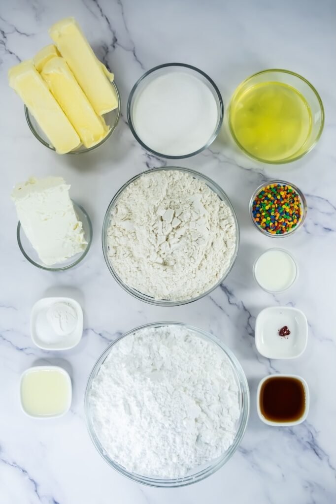 Flat lay of ingredients for Crumbl confetti cake cookies including butter, sugar, egg whites, flour, powdered sugar, cream cheese, vanilla, milk, baking powder, and colorful sequin sprinkles on a marble surface.