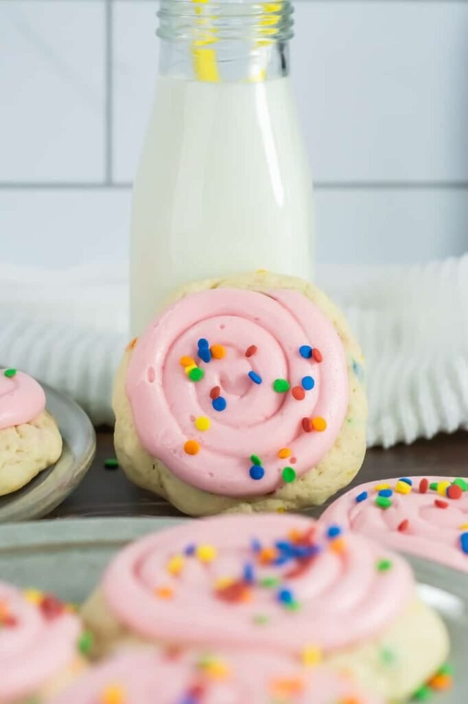 Close-up view of a confetti cake cookie topped with a smooth spiral of pink cream cheese frosting and colorful round sprinkles, positioned in front of a glass bottle filled with milk. The cookie is thick and pale in color with visible sprinkles baked into the dough, while additional frosted cookies sit on plates in the background on a wooden surface. The image creates a cozy dessert scene showing the finished cookies ready to enjoy with milk.