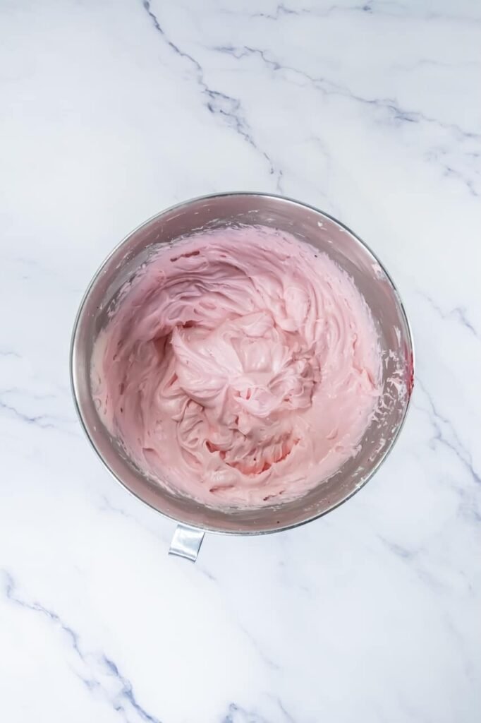 Overhead view of smooth, fluffy pink cream cheese frosting in a stand mixer bowl, showing a soft pastel pink color and creamy texture. This image shows the finished frosting after adding food coloring, ready to be transferred to a piping bag for decorating.