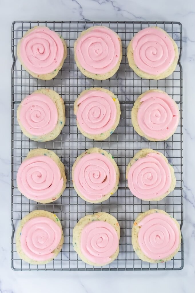 Overhead view of fully frosted confetti cake cookies cooling on a wire rack, each topped with a smooth spiral of pink cream cheese frosting. The cookies are thick, pale in color, and speckled with colorful sprinkles, showing the finished Crumbl-style cookies ready to serve.