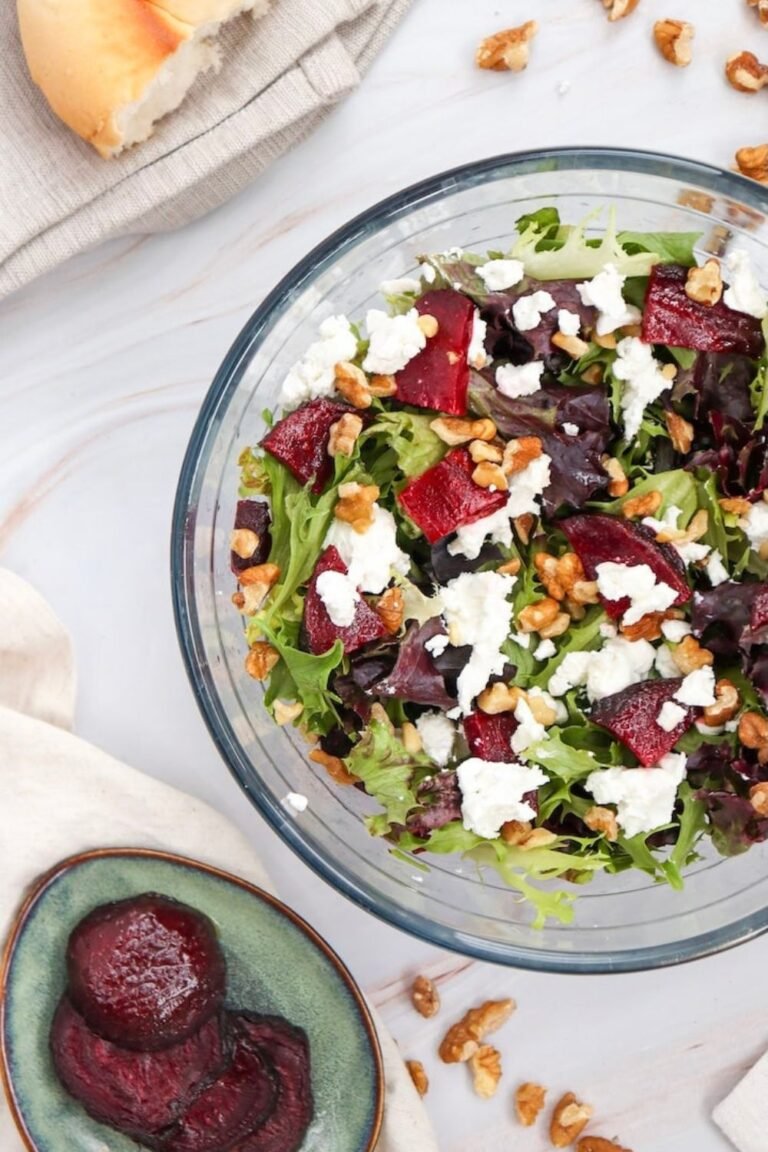 Finished beet goat cheese salad in a clear glass bowl made with mixed greens, roasted beet pieces, crumbled goat cheese, and chopped walnuts, styled on a light marble surface with extra roasted beets nearby.
