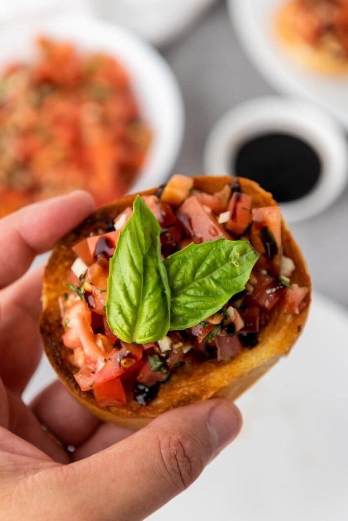 Close-up of a hand holding a slice of toasted bread topped with fresh tomato basil bruschetta and balsamic glaze, garnished with whole basil leaves, with more topping and glaze blurred in the background.