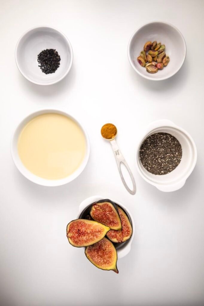 Overhead flat lay of fig chia pudding ingredients on a white background, including fresh halved figs, chia seeds, oat milk, cinnamon, pistachios, and black sesame seeds arranged in small bowls.