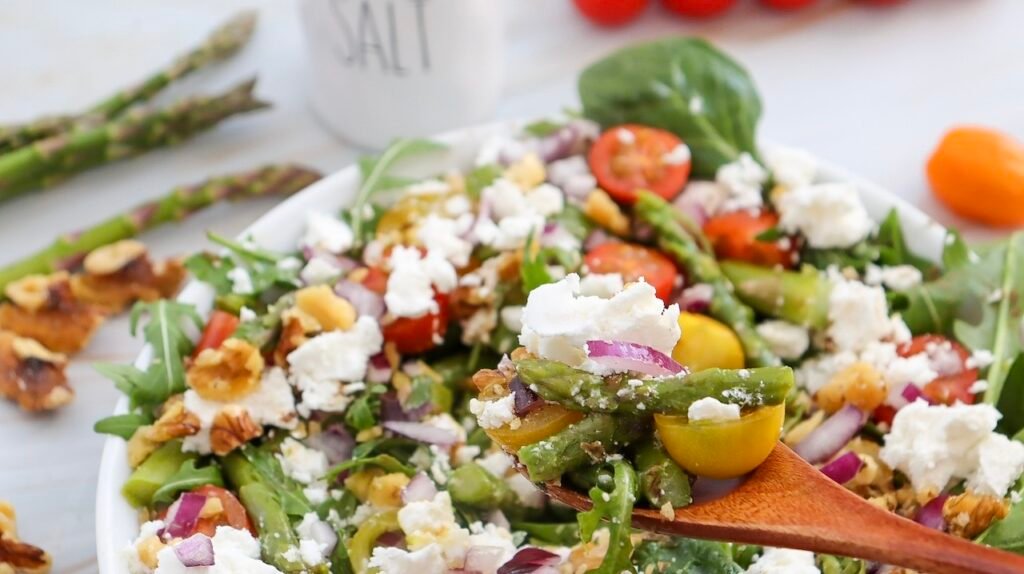 Angled close-up of a wooden spoon lifting a bite of spring goat cheese and asparagus salad from a bowl, showing tender asparagus, halved cherry tomatoes, red onion, walnuts, mixed greens, and crumbled goat cheese.