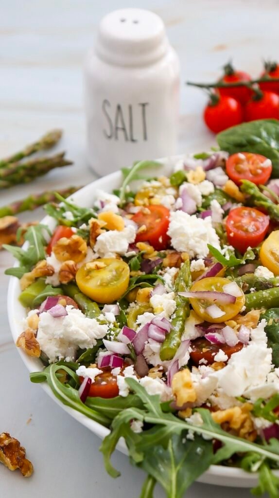 Angled close-up of a finished spring goat cheese and asparagus salad in a white bowl, featuring mixed greens, asparagus pieces, cherry tomatoes, red onion, walnuts, and crumbled goat cheese, with whole asparagus and a salt jar in the background.