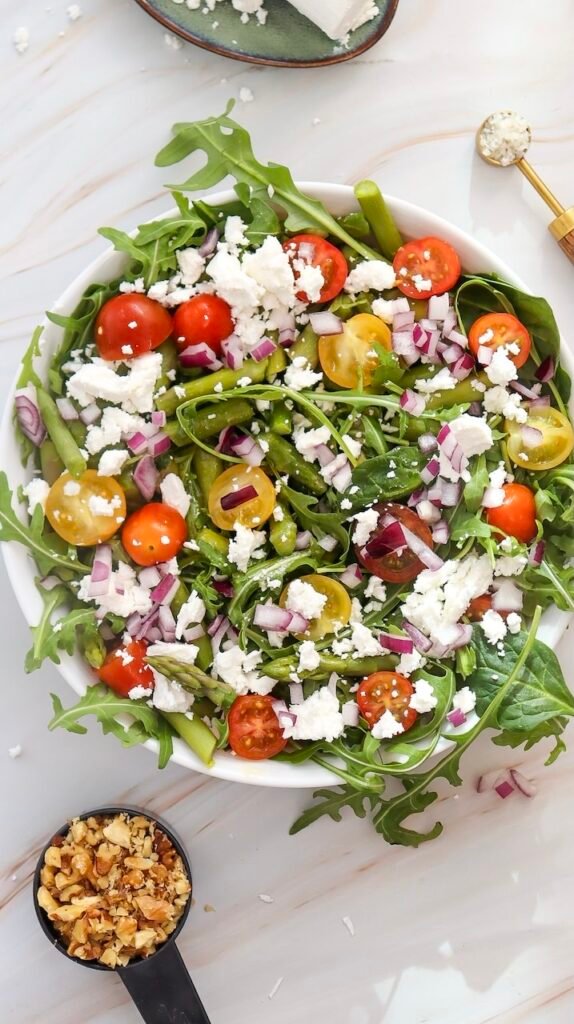 Overhead view of a finished goat cheese and asparagus salad with mixed greens, cherry tomatoes, red onion, walnuts, and crumbled goat cheese in a white bowl.