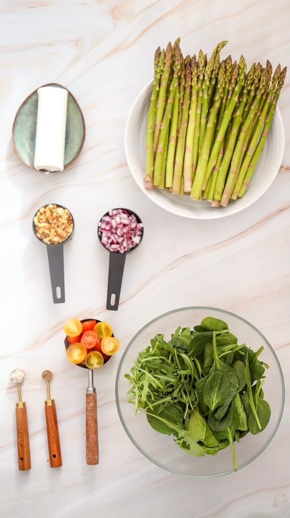 Overhead view of fresh ingredients for goat cheese and asparagus salad, including raw asparagus, mixed greens, goat cheese, cherry tomatoes, chopped red onion, and walnuts on a light countertop.