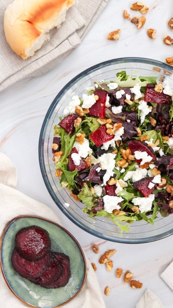Overhead view of beet goat cheese salad in a clear glass bowl made with mixed greens, roasted beet pieces, crumbled goat cheese, and chopped walnuts, styled on a light marble surface with extra roasted beets and bread nearby.