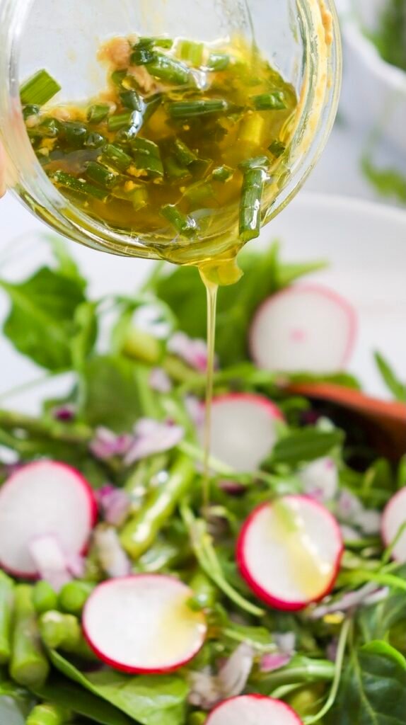Close-up of lemon Dijon dressing with chopped herbs being poured over a fresh asparagus and pea salad topped with sliced radishes and leafy greens.