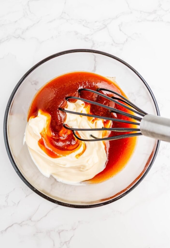 A whisk mixing mayonnaise and whiskey-flavored BBQ sauce in a glass bowl on a white marble surface, showing the process of making.