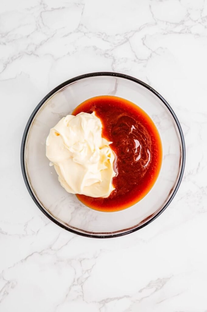 A glass bowl with mayonnaise and whiskey-flavored BBQ sauce side by side before mixing, showing the ingredients  on a white marble surface.