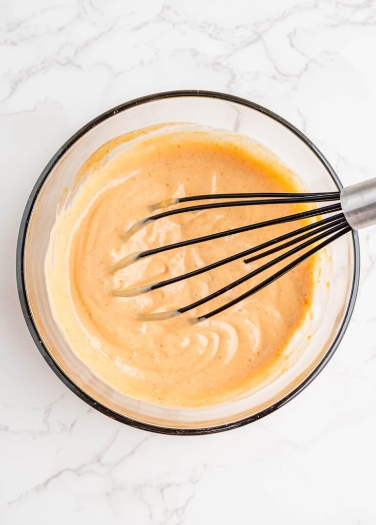 A whisk resting in a bowl of smooth, creamy after mixing mayonnaise and whiskey-flavored BBQ sauce on a white marble surface.