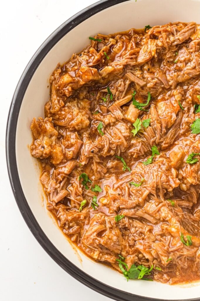 Bowl of tender shredded Instant Pot BBQ pulled pork in sauce, garnished with chopped parsley.
