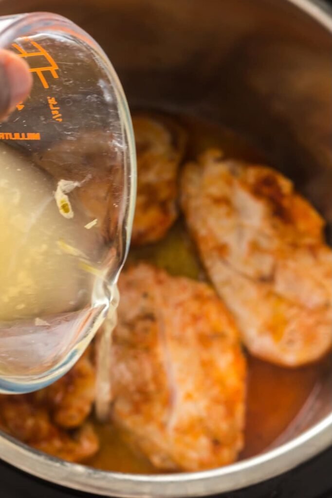 Chicken broth being poured from a glass measuring cup into an Instant Pot over browned, spice-rubbed pork shoulder steaks during the cooking process for Instant Pot pulled pork.