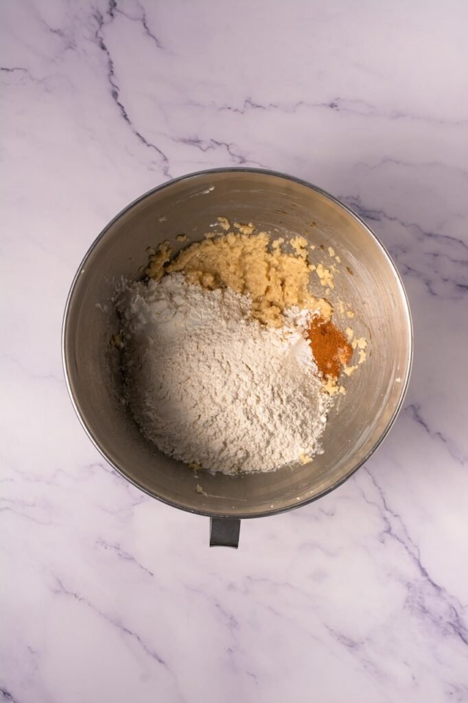 Overhead view of a metal stand mixer bowl on a marble countertop with creamed butter and sugar mixture topped with flour, cinnamon, cornstarch, baking powder, baking soda, and salt before mixing to form the dough.
