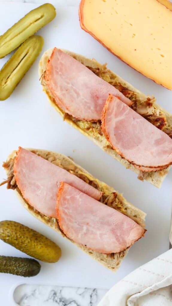 Overhead view of Cuban bread spread with mustard and layered with roast pork, topped with folded slices of ham during Cuban sandwich assembly, with pickles and Swiss cheese nearby on a cutting board.