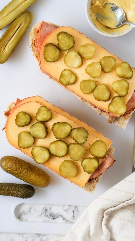 Overhead view of bread layered with roast pork, ham, and Swiss cheese, topped evenly with sliced dill pickles during assembly, with mustard and whole pickles nearby on a cutting board.