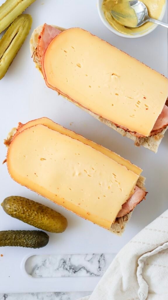 Overhead view of bread layered with roast pork and ham, topped with slices of Swiss cheese during assembly, with pickles and mustard nearby on a cutting board.