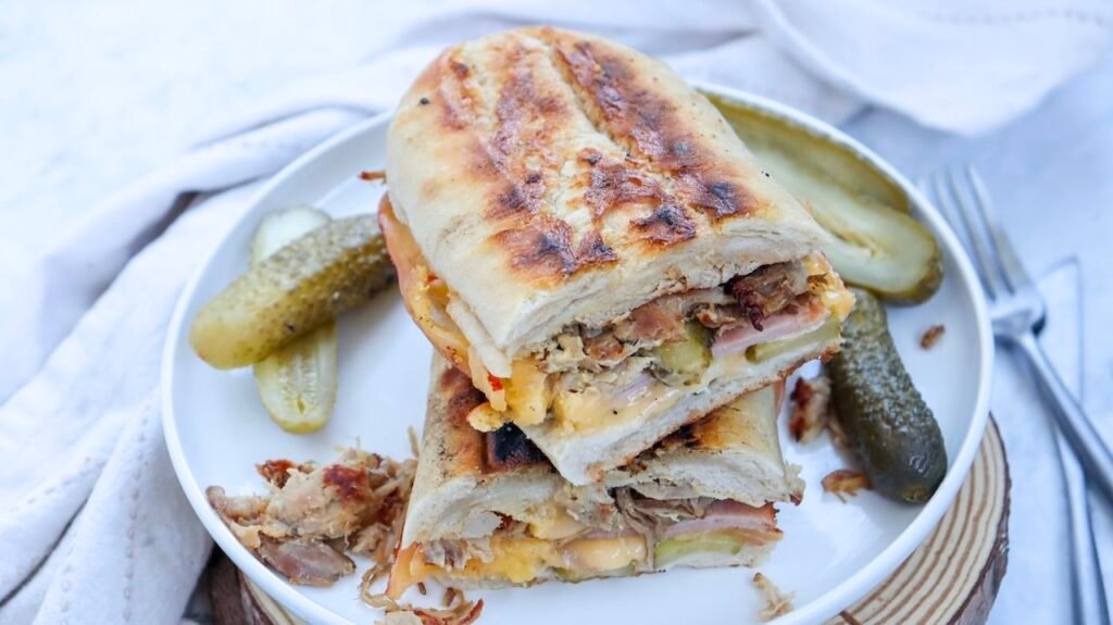 Stacked toasted Cuban sandwich halves on a white plate with pickles, showing layers of roast pork, ham, melted cheese, and sliced pickles inside crispy golden bread.