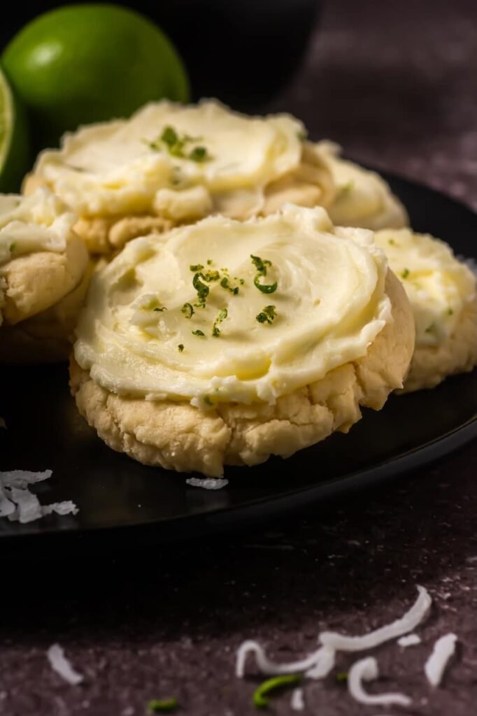 Close-up of thick coconut lime cookies topped with creamy lime buttercream frosting and fresh lime zest on a dark plate with whole limes in the background.