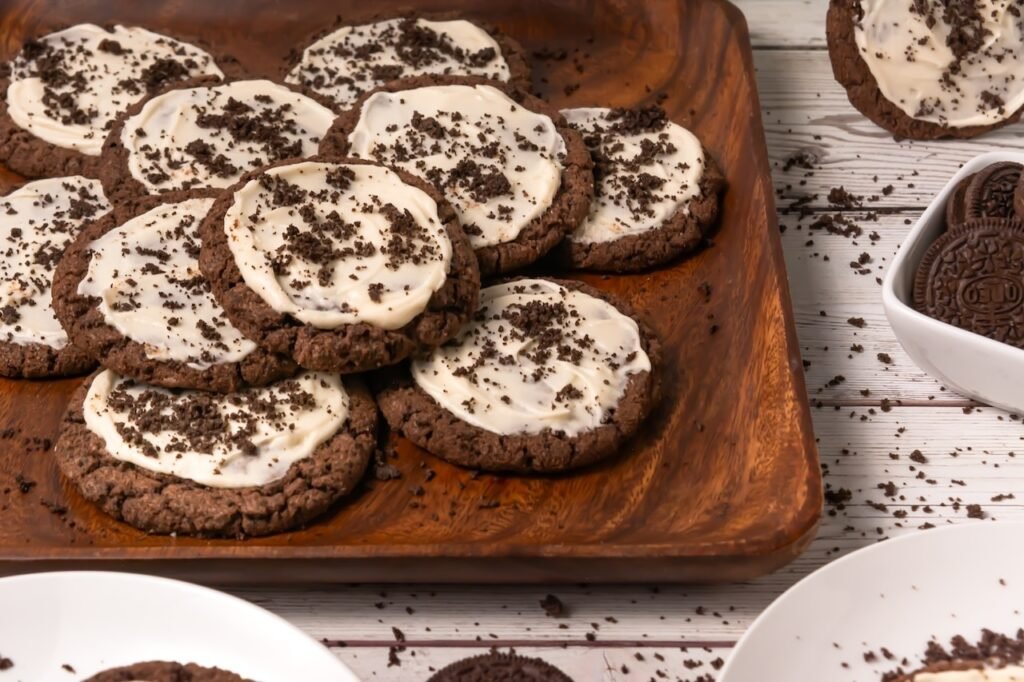 Close-up of crumbl chocolate Oreo cookies with frosting and Oreo crumbs arranged on a wooden serving tray, with additional cookies and crumbs in the background.