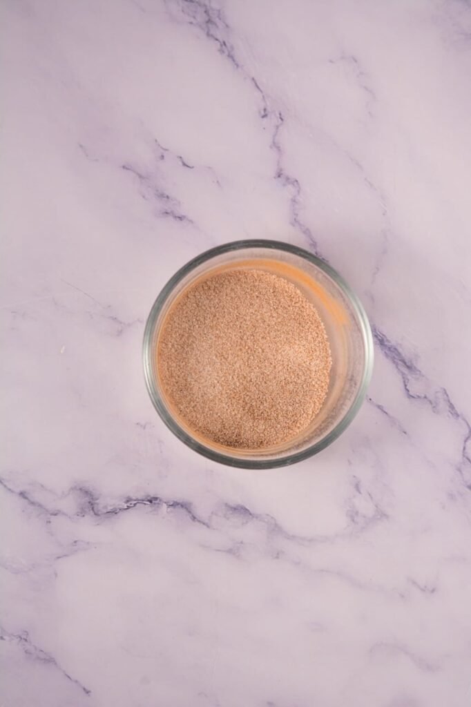 Overhead view of a small glass bowl filled with a cinnamon sugar mixture used as a finishing sprinkle for Crumbl cinnamon swirl cookies, sitting on a light marble countertop.