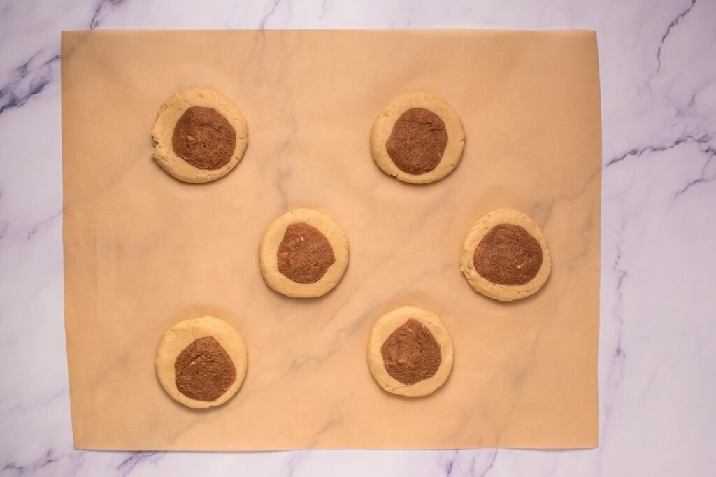 Overhead view of six scoops of cookie dough on parchment paper with a ball of cinnamon sugar topping pressed into the center of each piece of dough, prepared for baking Crumbl cinnamon swirl cookies.