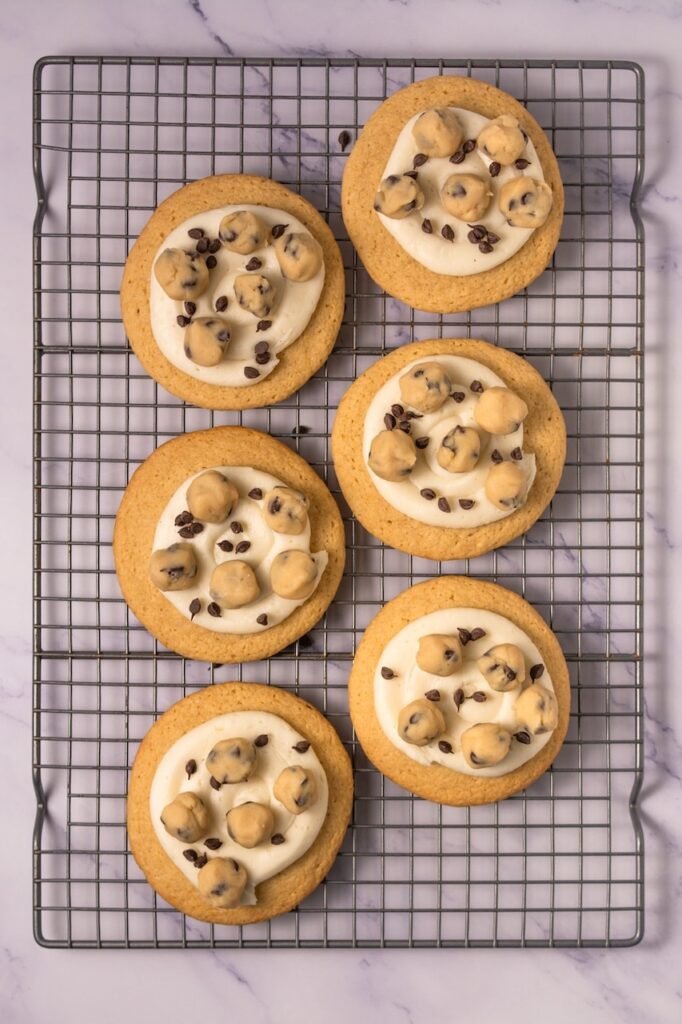 Homemade desserts on a wire cooling rack topped with creamy vanilla frosting, small balls of edible chocolate chip cookie dough, and a sprinkle of mini chocolate chips.