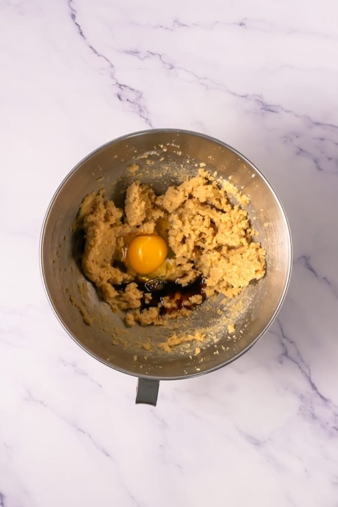 Stand mixer bowl containing creamed butter and sugars with a whole egg and vanilla added before mixing cookie dough.