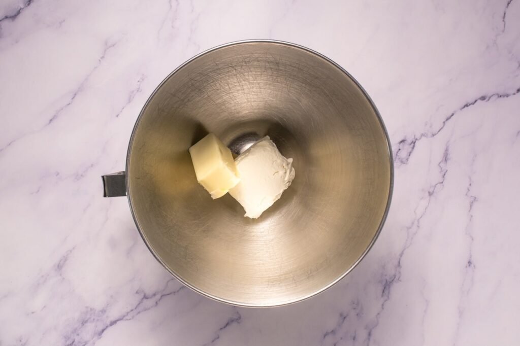 Overhead view of a metal stand mixer bowl on a marble countertop containing softened cream cheese and butter before mixing together to make the frosting for Crumbl cinnamon swirl cookies.