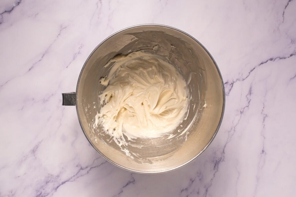 Overhead view of a metal stand mixer bowl on a marble countertop containing smooth cream cheese frosting after mixing butter, cream cheese, powdered sugar, and vanilla for Crumbl cinnamon swirl cookies.