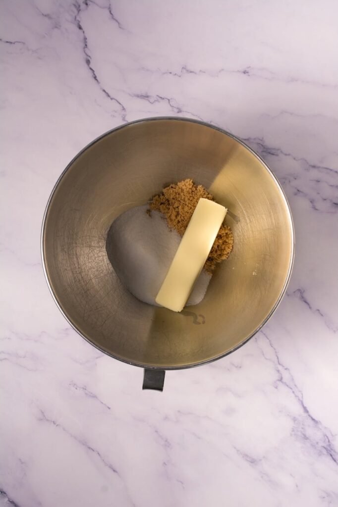 Overhead view of a metal stand mixer bowl on a light marble countertop containing softened butter, white granulated sugar, and brown sugar before creaming together to make the dough.