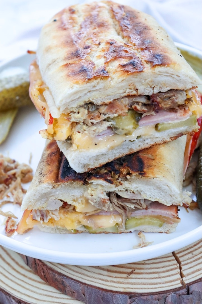 Close-up of a toasted Cuban sandwich cut into halves and stacked on a plate, showing layers of roast pork, ham, melted cheese, and pickles inside crispy golden bread.