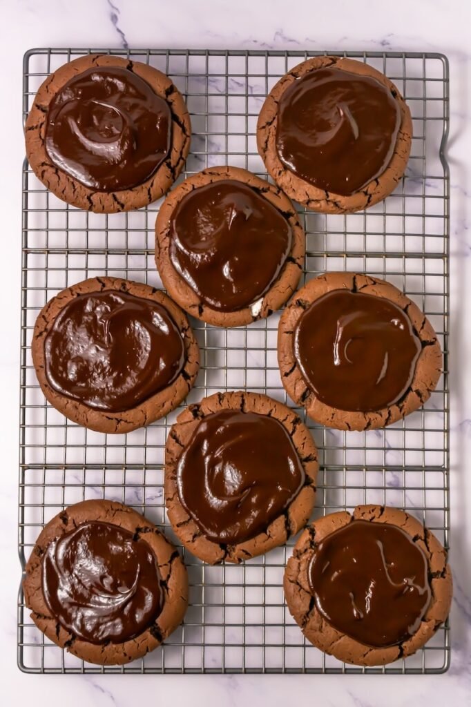 Chocolate mallow cupcake cookies on a cooling rack topped with smooth chocolate ganache before adding the white chocolate drizzle.