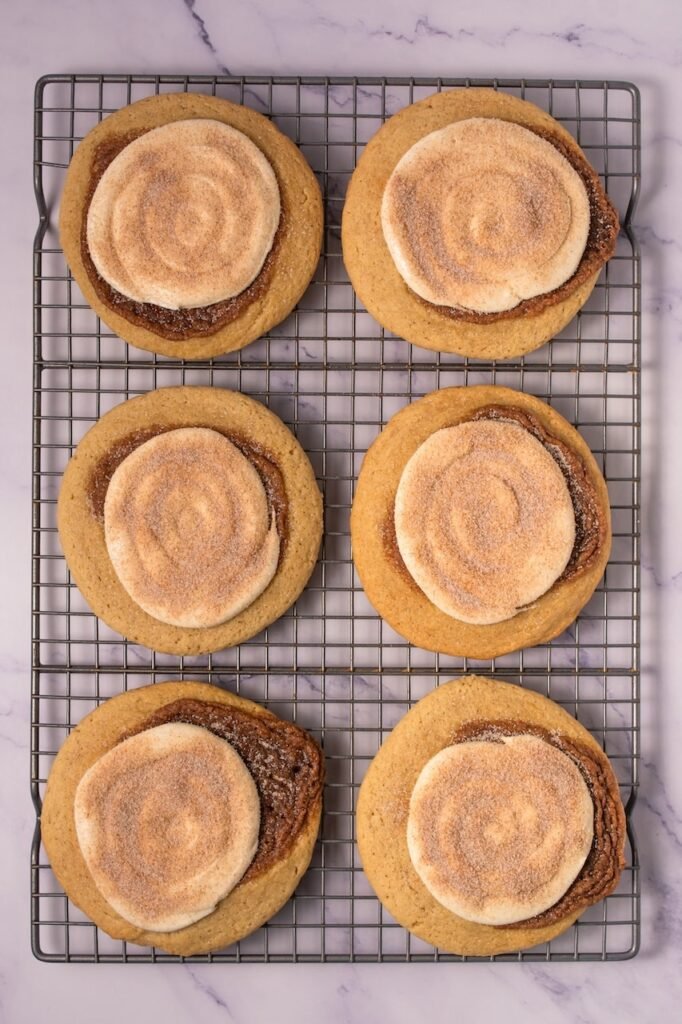 Overhead view of six baked Crumbl cinnamon swirl cookies on a wire cooling rack, each topped with cream cheese frosting and finished with a light cinnamon sugar sprinkle on top.