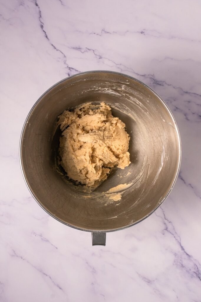 Overhead view of a metal stand mixer bowl on a marble countertop containing thick cookie dough after mixing butter, sugars, egg, vanilla, and dry ingredients to prepare the dough.
