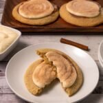 Overhead view of six baked Crumbl cinnamon swirl cookies on a wire cooling rack, each topped with cream cheese frosting and finished with a light cinnamon sugar sprinkle.