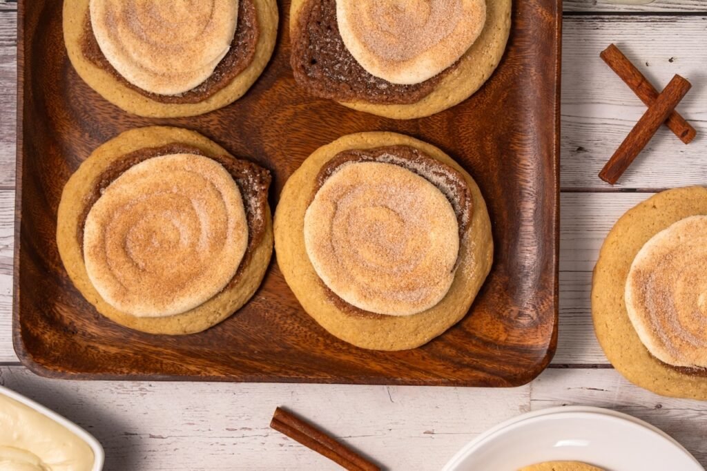 Overhead view of several Crumbl cinnamon swirl cookies arranged on a wooden serving tray with cinnamon sticks nearby, showing the thick cookie texture, cinnamon swirl layer, and cream cheese frosting topping.