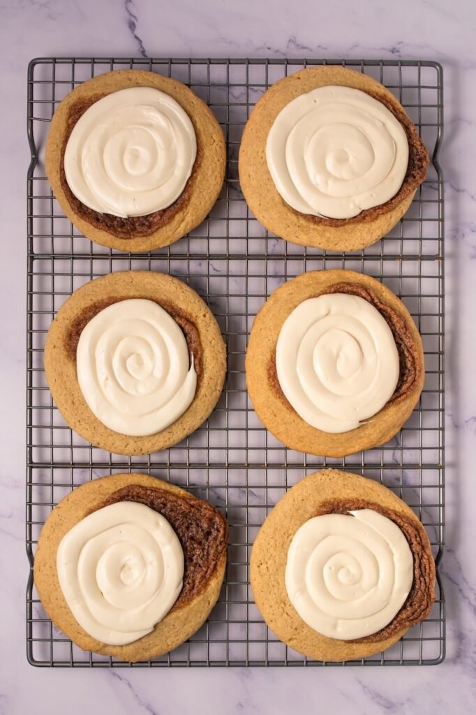 Overhead view of six baked Crumbl cinnamon swirl cookies on a wire cooling rack, each topped with a spiral of cream cheese frosting and showing the cinnamon swirl baked into the cookie.