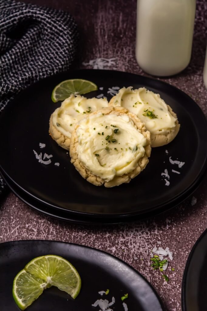 Three thick coconut lime cookies topped with creamy lime frosting and lime zest served on a black plate with lime slices and coconut flakes.