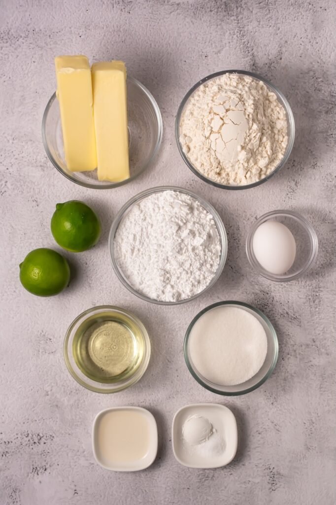 Overhead view of ingredients for coconut lime cookies including butter, flour, powdered sugar, granulated sugar, lime juice, fresh limes, egg, baking powder, and oil arranged in small bowls.